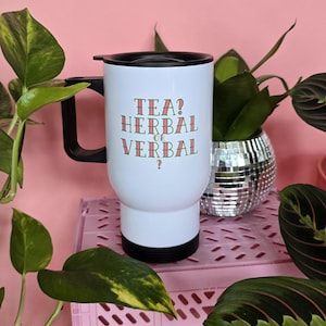 Op de afbeelding: Witte reistumbler met een zwarte deksel en handvat. De tumbler heeft de tekst "TEA? HERBAL OR VERBAL?" erop gedrukt in groen en roze.