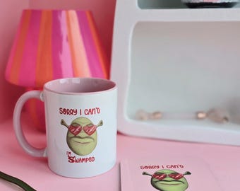 May include: A white ceramic mug with a pink interior and a matching square coaster. Both items feature a green ogre character wearing heart-shaped sunglasses with the text "SORRY I CAN'T I'M SWAMPED" printed in red and black lettering on the front.