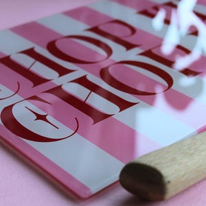 May include: A rectangular glass cutting board featuring alternating vertical stripes of light pink and white. The board displays the text CHOP SHOP in bold red capital letters with star accents. A wooden handle is attached to the side of the board.