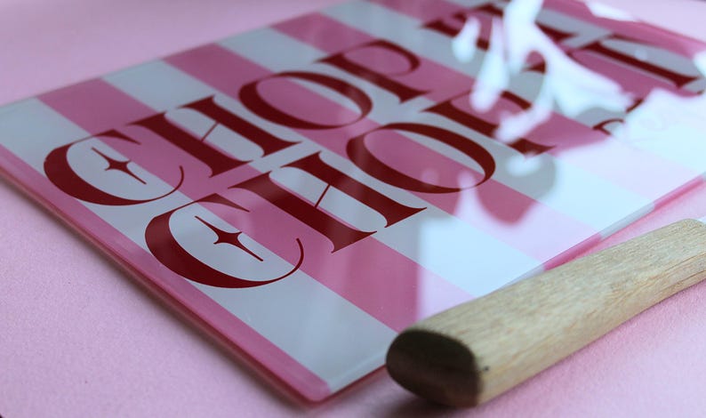 May include: A rectangular glass cutting board featuring alternating vertical stripes of light pink and white. The board displays the text CHOP SHOP in bold red capital letters with star accents. A wooden handle is attached to the side of the board.