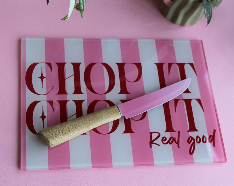 May include: A rectangular glass cutting board with vertical pink and white stripes and the red text CHOP IT CHOP IT Real good. A kitchen knife with a light wood handle and a pink blade is placed diagonally across the center of the board.