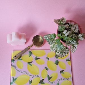 May include: A rectangular glass cutting board with a pink background and a yellow lemon pattern featuring the text WHEN LIFE GIVES YOU LEMONS MAKE LIMONCELLO, a brass measuring scoop with a wooden handle, a pink flower-shaped candle, and a plant.