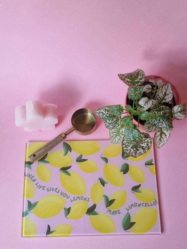 May include: A rectangular glass cutting board with a pink background and a yellow lemon pattern featuring the text WHEN LIFE GIVES YOU LEMONS MAKE LIMONCELLO, a brass measuring scoop with a wooden handle, a pink flower-shaped candle, and a plant.