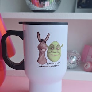 May include: White travel mug with a black handle and lid. The mug features illustrations of Shrek and Donkey with the text "Shrek runs on coffee, Donkey runs on conversation."