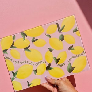 May include: A rectangular glass cutting board featuring a repeating pattern of yellow lemons with green leaves on a light pink background. The text "WHEN LIFE GIVES YOU LEMONS MAKE LIMONCELLO!" is printed in green letters across the center surface.