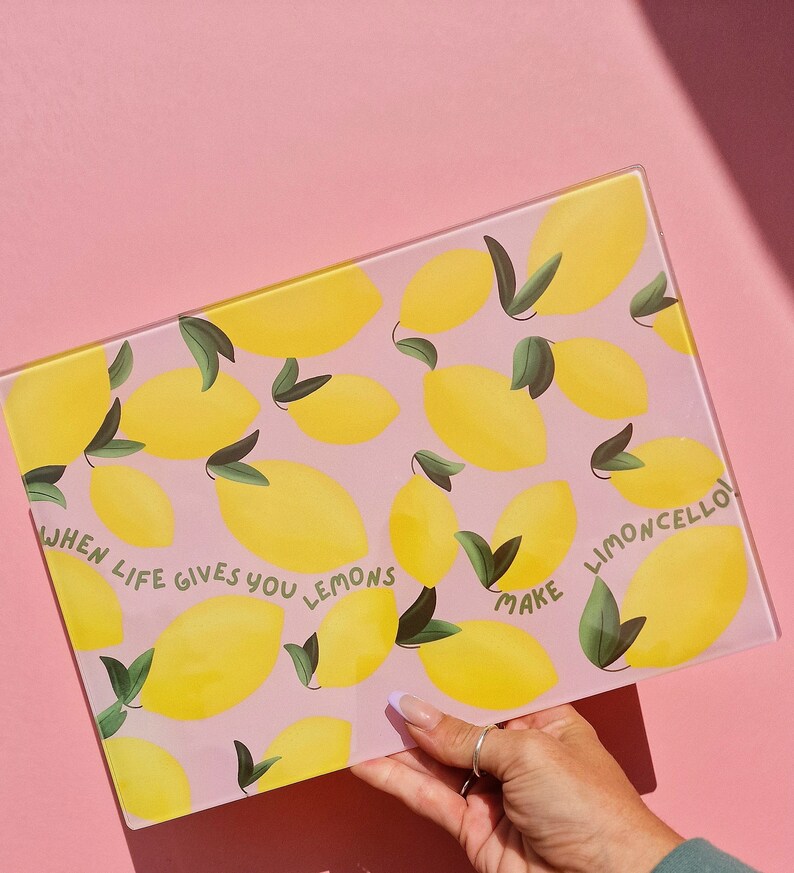 May include: A rectangular glass cutting board featuring a repeating pattern of yellow lemons with green leaves on a light pink background. The text "WHEN LIFE GIVES YOU LEMONS MAKE LIMONCELLO!" is printed in green letters across the center surface.