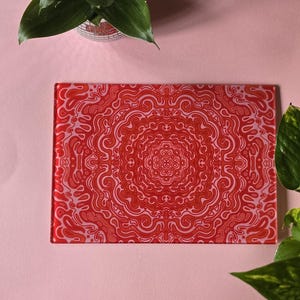 Puede incluir: Una tabla de cortar de vidrio roja con un diseño de mandala rosa y naranja. La tabla está sobre una superficie rosa.