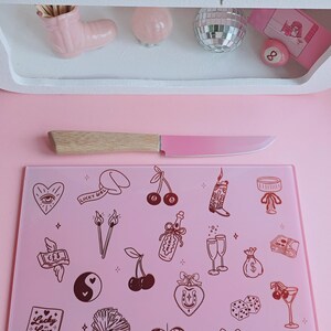 May include: A rectangular light pink glass cutting board featuring a repeating pattern of dark red line art illustrations including cherries, a disco ball, dice, a horseshoe, a hand holding cash, and a matchbox labeled Lucky in Love.