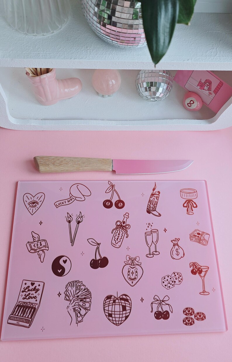 May include: A rectangular light pink glass cutting board featuring a repeating pattern of dark red line art illustrations including cherries, a disco ball, dice, a horseshoe, a hand holding cash, and a matchbox labeled Lucky in Love.