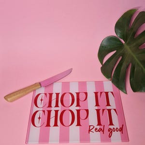 May include: A rectangular glass cutting board with a pink and white vertical striped pattern. The text "CHOP IT CHOP IT Real good" is printed in red. A knife with a pink blade and a light brown wooden handle rests on the board.