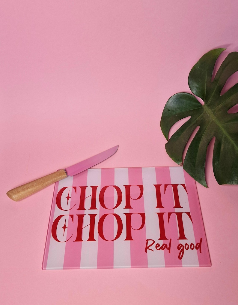 May include: A rectangular glass cutting board with a pink and white vertical striped pattern. The text "CHOP IT CHOP IT Real good" is printed in red. A knife with a pink blade and a light brown wooden handle rests on the board.