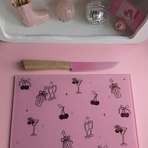 Puede incluir: Una tabla de cortar de vidrio rosa con ilustraciones negras de cerezas, botellas de champán y copas de cóctel. Un cuchillo rosa con mango de madera descansa sobre la tabla. Una estantería superior contiene objetos decorativos, incluyendo una bola de discoteca.