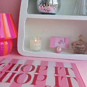 May include: A rectangular glass cutting board with pink and white stripes and the text "CHOP IT CHOP IT Real good" in red. A white arched shelf displays a disco ball planter with a green plant, a candle, a pink card, a pink number 8 ball, and a glass jar.