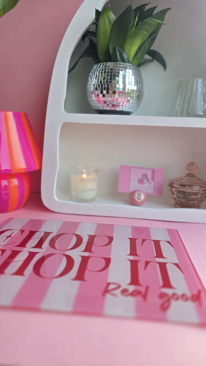 May include: A rectangular glass cutting board with pink and white stripes and the text "CHOP IT CHOP IT Real good" in red. A white arched shelf displays a disco ball planter with a green plant, a candle, a pink card, a pink number 8 ball, and a glass jar.