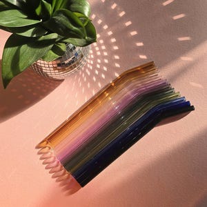Puede incluir: Un juego de 10 pajitas para beber de vidrio en varios colores, incluyendo ámbar, morado, verde y azul. Las pajitas están dobladas y dispuestas en un patrón de arcoíris.