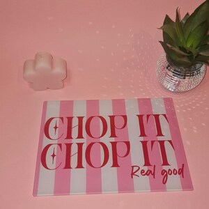 May include: A rectangular glass cutting board featuring vertical pink and white stripes with the red text CHOP IT CHOP IT Real good printed on the surface. The board is positioned on a pink surface next to a flower-shaped candle and a plant.