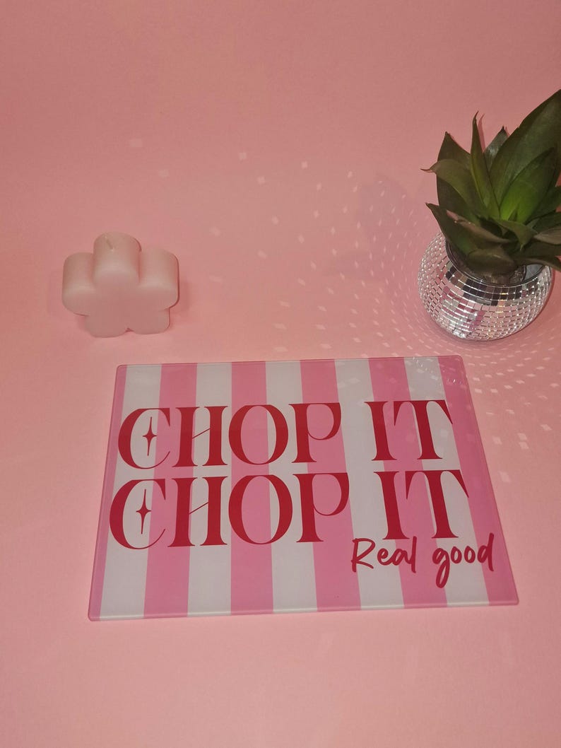 May include: A rectangular glass cutting board featuring vertical pink and white stripes with the red text CHOP IT CHOP IT Real good printed on the surface. The board is positioned on a pink surface next to a flower-shaped candle and a plant.