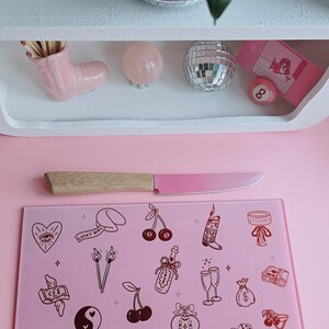 May include: A rectangular pink glass cutting board featuring a variety of dark brown line art illustrations including cherries, dice, a disco ball, a cowboy boot, and a matchbox labeled Lucky Love, paired with a knife with a light wood handle and pink blade.