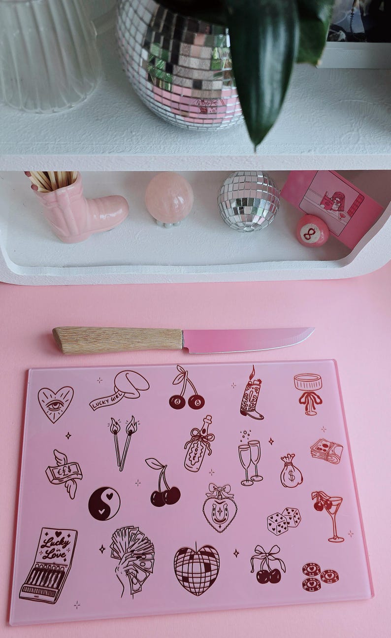 May include: A rectangular pink glass cutting board featuring a variety of dark brown line art illustrations including cherries, dice, a disco ball, a cowboy boot, and a matchbox labeled Lucky Love, paired with a knife with a light wood handle and pink blade.