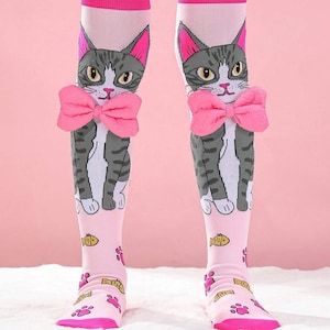 Könnte beinhalten: Ein Paar rosa Kniestrümpfe mit einem grau-weißen Katzenmotiv. Die Katze trägt eine rosa Schleife und die Socken haben rosa Pfotenabdrücke und Fischmotive.