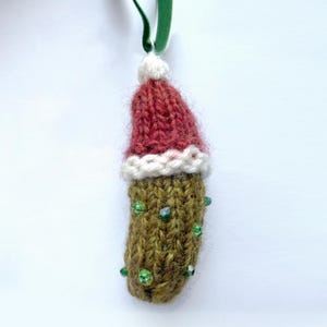 May include: A handmade Christmas ornament shaped like a pickle, with a red knit Santa hat and green beaded embellishments. The ornament is suspended by a green velvet ribbon, perfect for holiday decor.