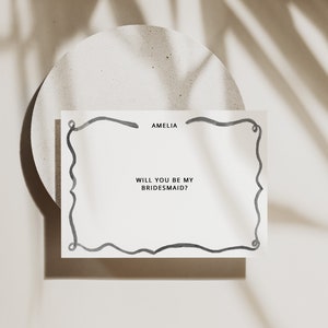 以下が含まれることがあります： アメリアと「私のブライズメイドになってくれますか？」というテキストが書かれた、黒い手書きの枠のある白いカード
