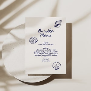 Pode incluir: Um cartão de menu branco com ilustrações de conchas marinhas em tinta azul. O texto diz "On The Menu" e lista os itens do menu: "Start: azeitonas marinadas", "Share: focaccia e azeite de oliva, rotolo de espinafre e pinhões com rúcula e parmesão, salada panzanella defumada", "Finish: tiramisu".