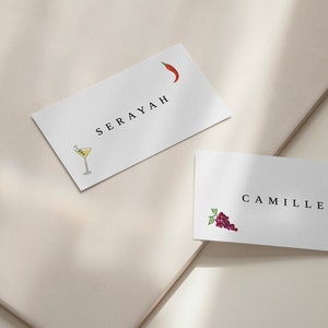 Op de afbeelding: Twee witte placecards met zwarte tekst en illustraties. De bovenste kaart zegt "SERAYAH" en heeft een rode peperillustratie. De onderste kaart zegt "CAMILLE" en heeft een paarse druiventros en een rode kreeft-illustratie.