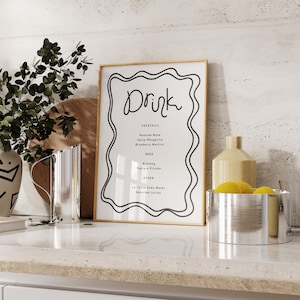 Pode incluir: Uma impressão emoldurada com uma moldura dourada e uma ilustração em preto e branco de um menu de bebidas. O menu inclui coquetéis, cerveja e outras bebidas. A impressão está sobre uma bancada com uma tigela de prata cheia de limões e um vaso com uma garrafa de cerâmica amarela.