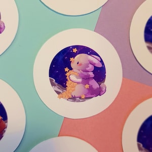 May include: A purple bunny with pink ears holds a pile of gold stars in front of a dark blue galaxy background with white stars. The bunny is sitting on a white, round sticker.