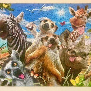 May include: A colorful jigsaw puzzle featuring a zebra, a giraffe, a rhino, a lemur, and a meerkat. The animals are all smiling and have their tongues sticking out. The puzzle is set against a blue sky with white clouds.