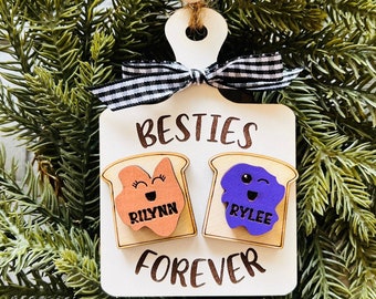 Pode incluir: Um ornamento de madeira branco em forma de tábua de cortar com uma fita xadrez preta e branca. Diz "Besties Forever" com duas fatias de torradas de desenho animado, uma com manteiga de amendoim e outra com geléia. A torrada de manteiga de amendoim diz "Rilynn" e a torrada de geléia diz "Rylee".