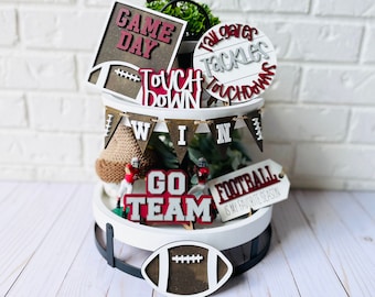 May include: A three-tiered white tiered tray decorated with football-themed wooden signs. The signs say "Game Day", "Touch Down", "Tackles Touchdowns", "Win", "Go Team", and "Football is my favorite season".