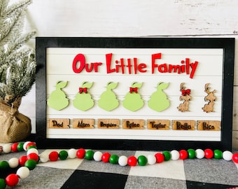 May include: A framed wooden sign with the words "Our Little Family" in red letters. The sign features green and brown wooden figures with red bows and the names "Dad", "Mom", "Brayden", "Rylee", "Taylor", "Bella", and "Rico" written on wooden plaques.
