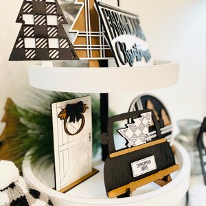 May include: A tiered tray decorated with black and white checkered Christmas decorations. The tray features a black truck with a plaid Christmas tree in the back, a black and white door with a wreath, a black and white plaid Christmas tree, and a sign that reads "From Our Christmas".