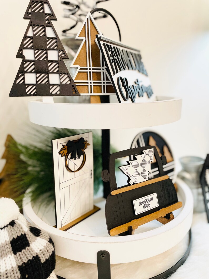 May include: A tiered tray decorated with black and white checkered Christmas decorations. The tray features a black truck with a plaid Christmas tree in the back, a black and white door with a wreath, a black and white plaid Christmas tree, and a sign that reads "From Our Christmas".