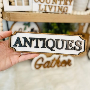 May include: A wooden sign with a white background and black lettering that reads "ANTIQUES". The sign has a brown wood frame and is slightly distressed.