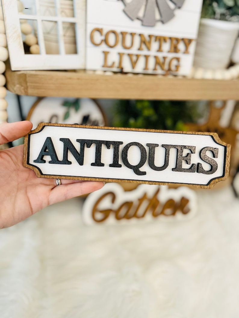 May include: A wooden sign with a white background and black lettering that reads "ANTIQUES". The sign has a brown wood frame and is slightly distressed.