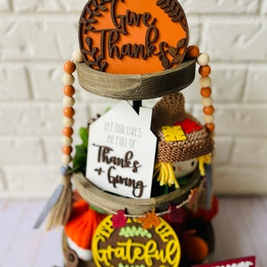 Może przedstawiać: Tr&oacute;jpoziomowa drewniana taca ozdobiona dekoracjami jesiennymi. G&oacute;rny poziom zawiera ozdobę w kształcie dyni z napisem "Give Thanks". Środkowy poziom ma ozdobę z napisem "Let our lives be full of Thanks + Giving" oraz małą figurkę straszyka. Dolny poziom zawiera ozdobę z napisem "The Grateful Blessed" oraz ozdobę z napisem "Everyone Gather Together".