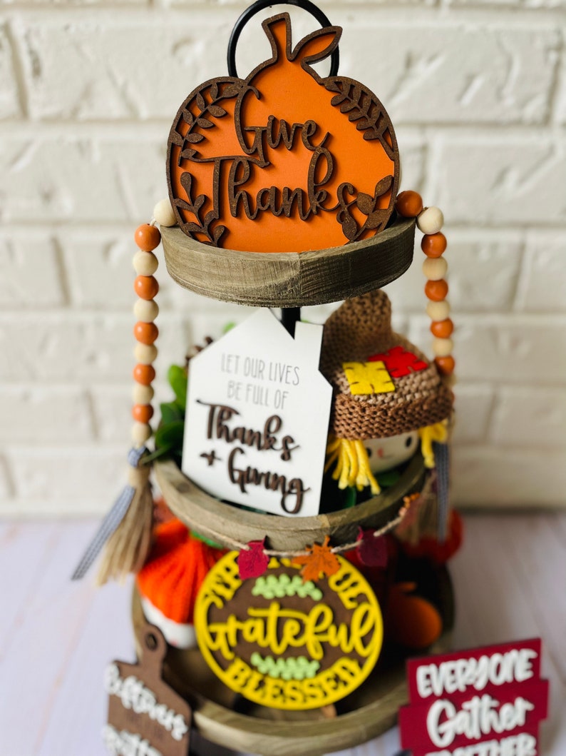 Może przedstawiać: Tr&oacute;jpoziomowa drewniana taca ozdobiona dekoracjami jesiennymi. G&oacute;rny poziom zawiera ozdobę w kształcie dyni z napisem "Give Thanks". Środkowy poziom ma ozdobę z napisem "Let our lives be full of Thanks + Giving" oraz małą figurkę straszyka. Dolny poziom zawiera ozdobę z napisem "The Grateful Blessed" oraz ozdobę z napisem "Everyone Gather Together".