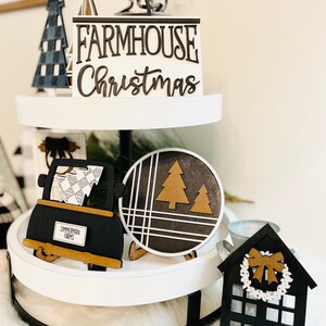 May include: A two-tiered white tiered tray with black metal accents. The top tier features a black and white checkered Christmas tree, a black and white sign that reads "FARMHOUSE Christmas", and a black and white spiral. The bottom tier features a black truck with a white and black checkered design, a black and white round sign with a brown tree, a small black house with a white wreath and a black bow, and a silver metal bucket.