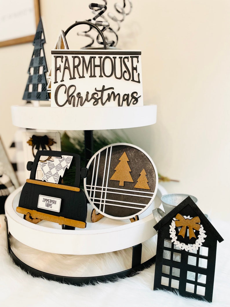 May include: A two-tiered white tiered tray with black metal accents. The top tier features a black and white checkered Christmas tree, a black and white sign that reads "FARMHOUSE Christmas", and a black and white spiral. The bottom tier features a black truck with a white and black checkered design, a black and white round sign with a brown tree, a small black house with a white wreath and a black bow, and a silver metal bucket.
