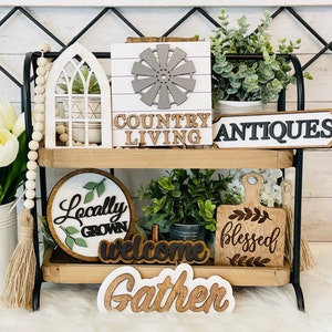 May include: A two-tiered wooden shelf with various farmhouse-style decor, including signs that say "Country Living", "Antiques", "Locally Grown", "Welcome", "Blessed", and "Gather".