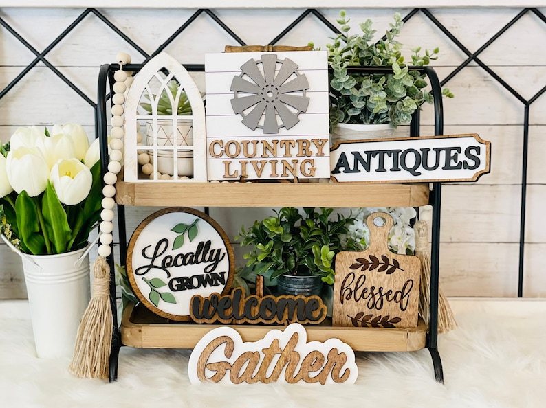 May include: A two-tiered wooden shelf with various farmhouse-style decor, including signs that say "Country Living", "Antiques", "Locally Grown", "Welcome", "Blessed", and "Gather".