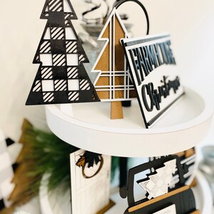 May include: A three-tiered white tiered tray decorated with black and white Christmas ornaments. The ornaments include a black and white plaid Christmas tree, a brown and white plaid Christmas tree, a black and white truck with a Christmas tree in the back, and a black and white sign that says "Farmhouse Christmas".