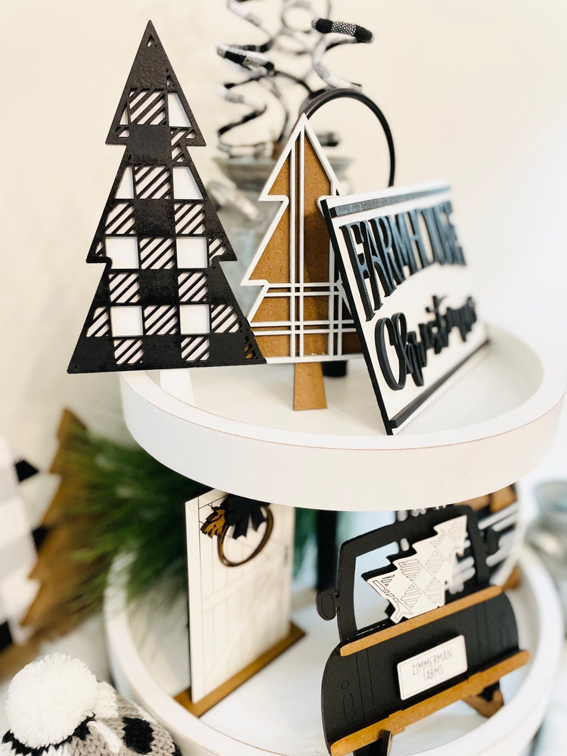 May include: A three-tiered white tiered tray decorated with black and white Christmas ornaments. The ornaments include a black and white plaid Christmas tree, a brown and white plaid Christmas tree, a black and white truck with a Christmas tree in the back, and a black and white sign that says "Farmhouse Christmas".