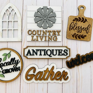 May include: A collection of eight wooden signs with various sayings and designs. The signs feature phrases like "Country Living", "Blessed", "Welcome", "Locally Grown", "Antiques", and "Gather".  One sign is a white silhouette of a window. Another sign features a gray windmill design.