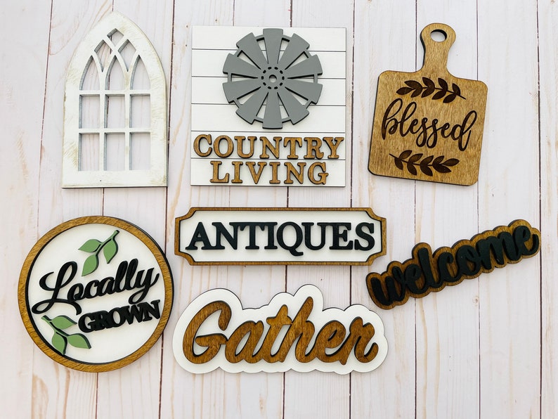 May include: A collection of eight wooden signs with various sayings and designs. The signs feature phrases like "Country Living", "Blessed", "Welcome", "Locally Grown", "Antiques", and "Gather".  One sign is a white silhouette of a window. Another sign features a gray windmill design.