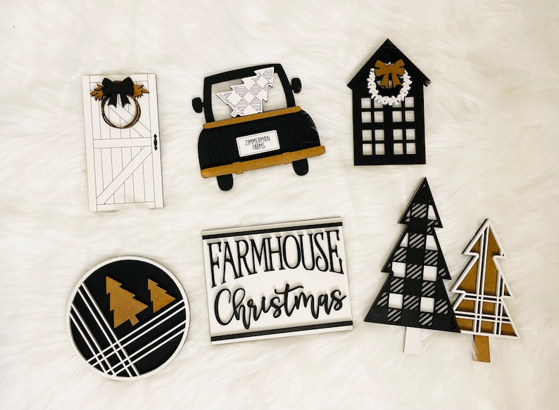 May include: A set of six black and white farmhouse Christmas decorations. The decorations include a truck with a Christmas tree in the back, a house with a wreath on the door, a door with a wreath, a round sign with two Christmas trees, and two Christmas trees with a black and white plaid pattern.