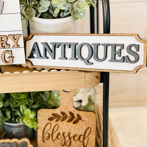 May include: A wooden sign with the word "ANTIQUE" in black letters on a white background. The sign is on a tiered wooden shelf with greenery and other decorative items.